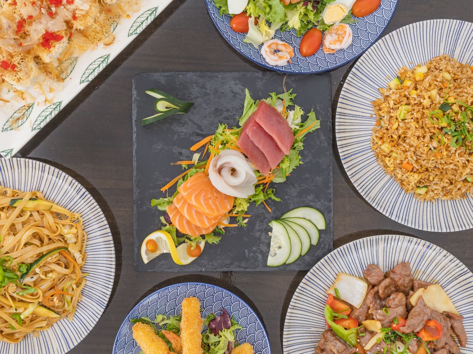 Overhead view of multiple Asian dishes including sashimi, fried rice, noodles, and beef